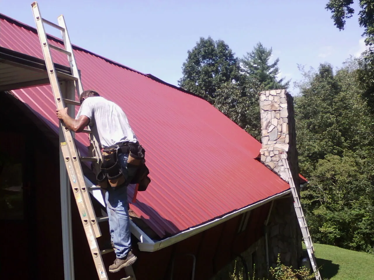 Roofer on a ladder beside a red metal roof and stone chimney — Soffit & Fascia Repair by Roofing Brothers in White Bird